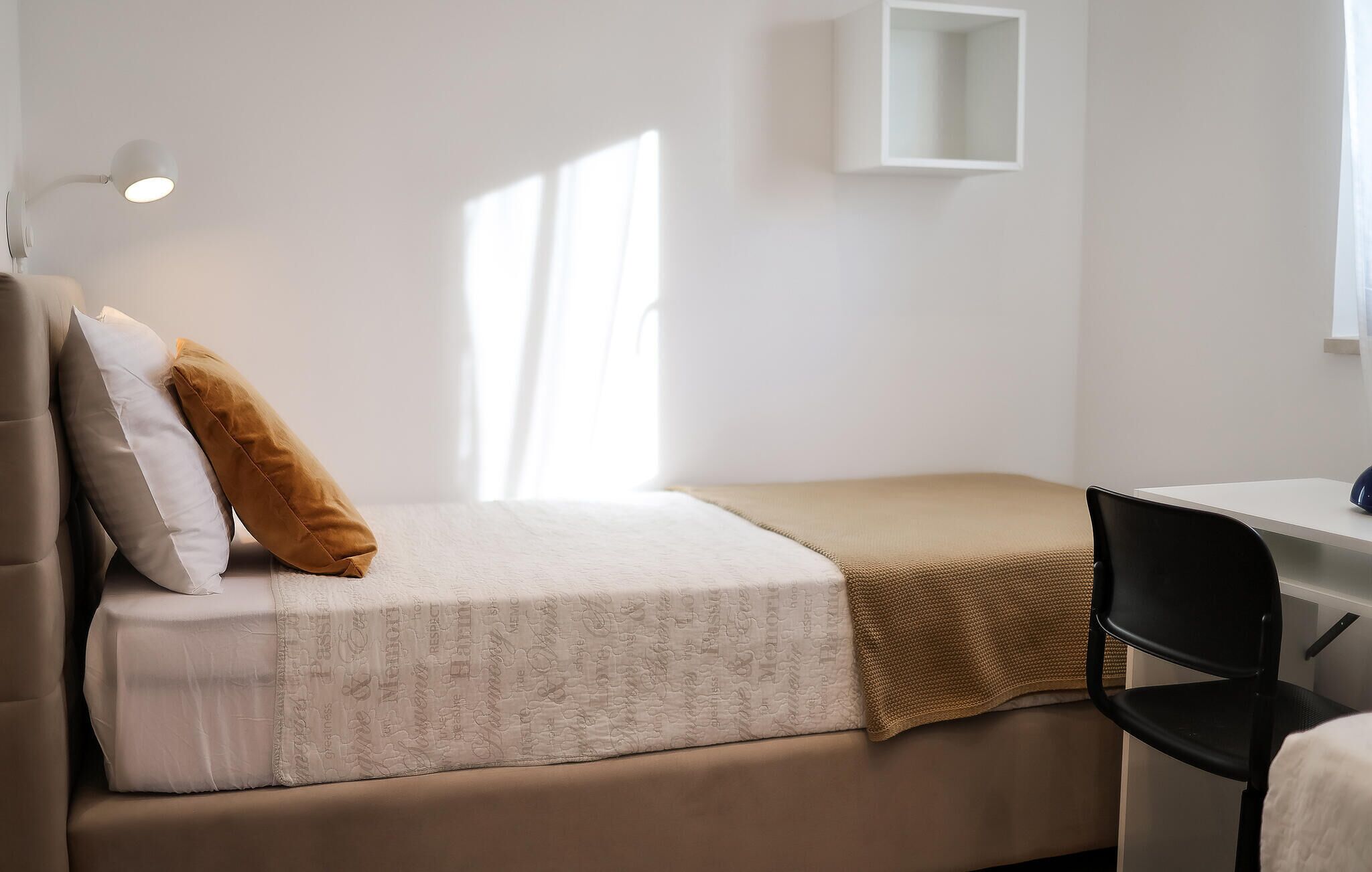Un lit simple avec une couvre-lit à motifs, un plaid marron et deux oreillers. Il y a une petite étagère blanche au mur et un bureau avec une chaise.