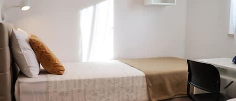 A single bed with a patterned bedspread, a brown throw blanket, and two pillows. There is a small white shelf on the wall and a desk with a chair.
