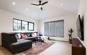 Ein modernes Wohnzimmer mit einer Eckcouch, einem Deckenventilator, großen Fenstern mit Jalousien und einem flachbildfernseher an der Wand.