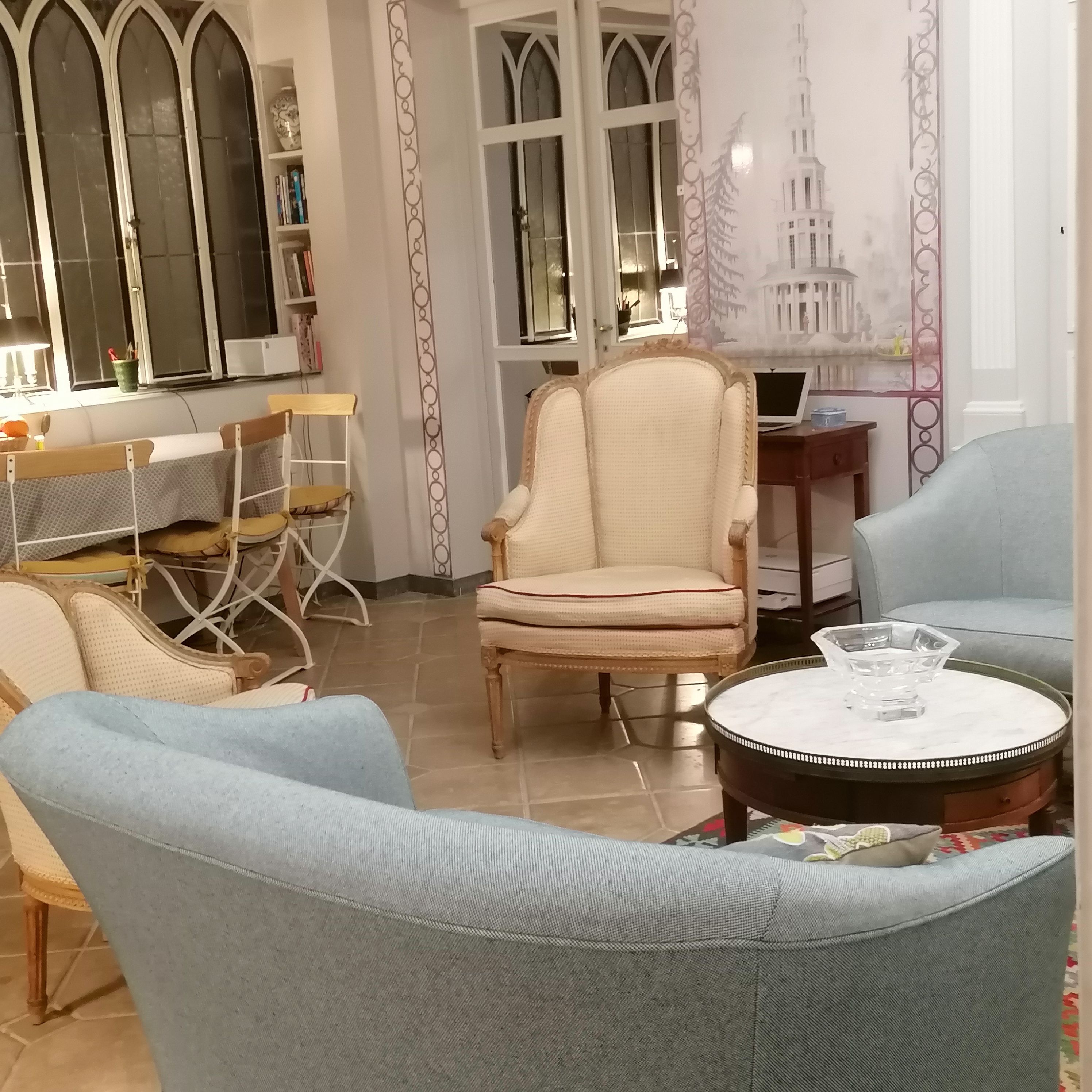 Een kamer met een ronde tafel, twee fauteuils, een boekenkast en een raam met decoratieve details.