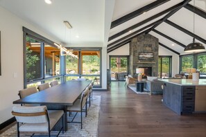 Une salle à manger spacieuse avec une longue table et des chaises, une cuisine dotée d’une cheminée en pierre et de grandes fenêtres offrant une vue sur l’extérieur.