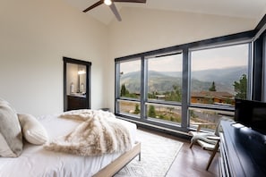 A bedroom with a large window, a bed, a dresser, a TV, and a ceiling fan. - Ultra-Luxury Mountain Retreat w/ Sauna & Hot Tub | Summit Sky Ranch (Silverthorne)