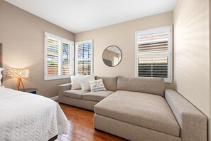 Ein Schlafzimmer mit einem Bett, zwei Sofas, einem runden Spiegel und Fenstern mit Jalousien.
