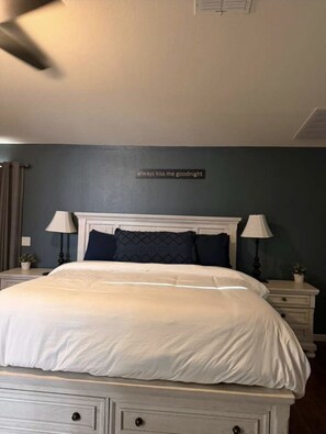 A bedroom with a bed, two bedside tables, a ceiling fan, and a wall with a sign. - Saguara Sunset: Tranquil Private Pool Oasis (Maricopa)