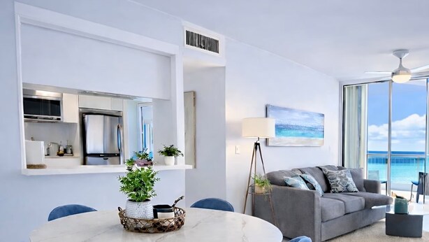 Una sala de estar moderna con una mesa de comedor redonda, sillas azules, un sofá gris y un ventanal con vista al océano.