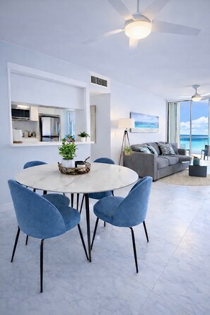 Un salon moderne avec une table à manger ronde, des chaises bleues, un canapé gris et une grande fenêtre donnant sur l’océan.
