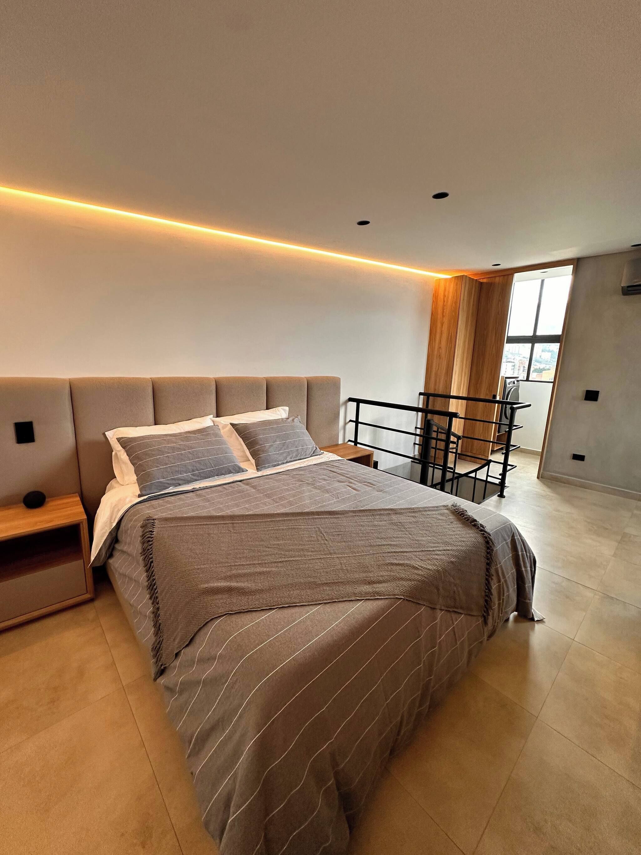 A modern bedroom with a large bed, a nightstand, a desk, and a window with a view.