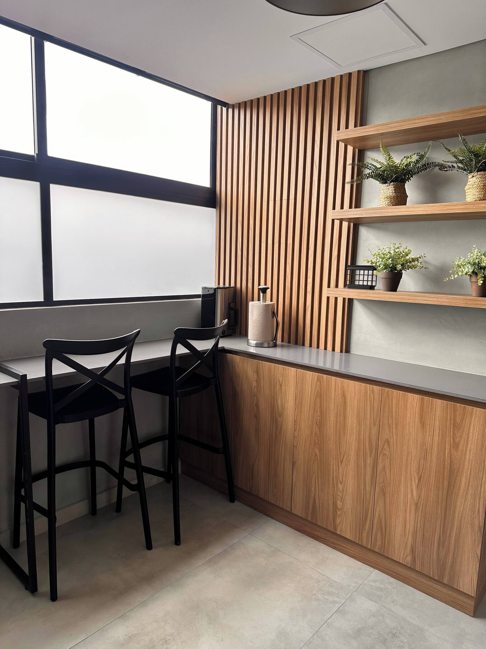 A modern kitchen with a wooden countertop, two black chairs, a wooden slatted wall, and a window.
