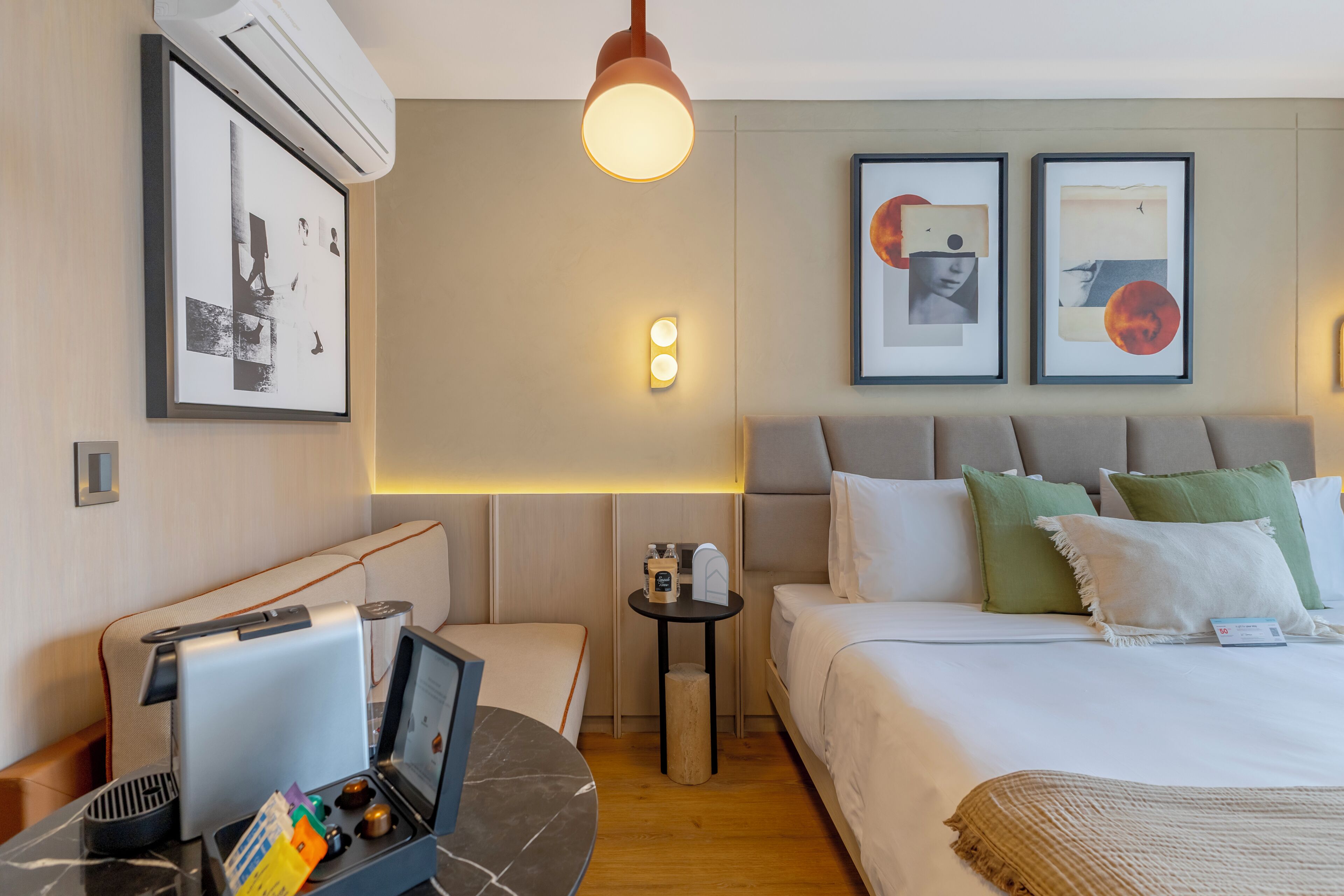 A hotel room with a bed, bedside table, lamp, wall art, and an air conditioning unit.