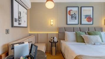 A hotel room with a bed, bedside table, lamp, wall art, and an air conditioning unit.