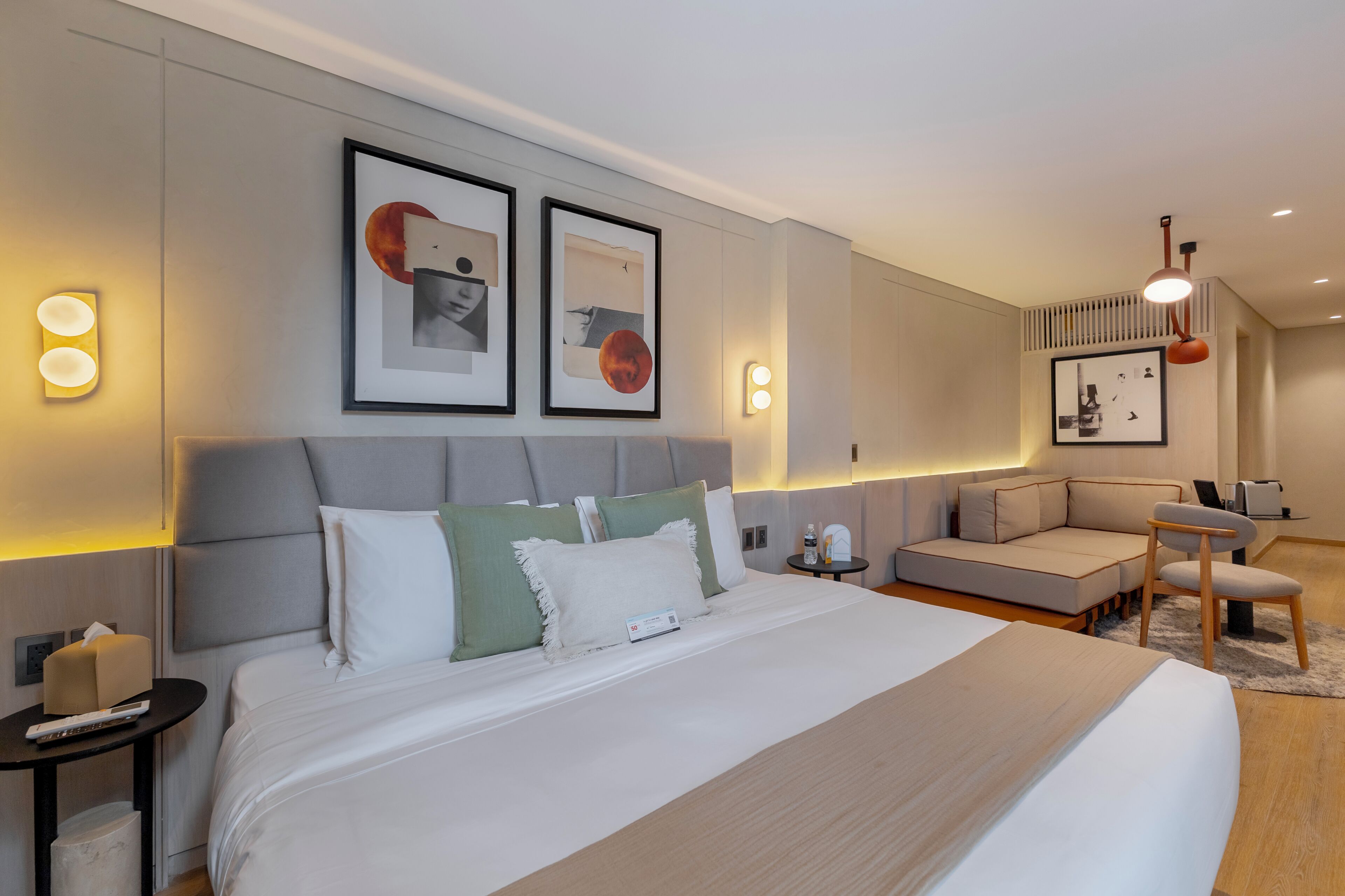 A modern hotel room with a large bed, a sofa, a small table, and wall-mounted light fixtures.