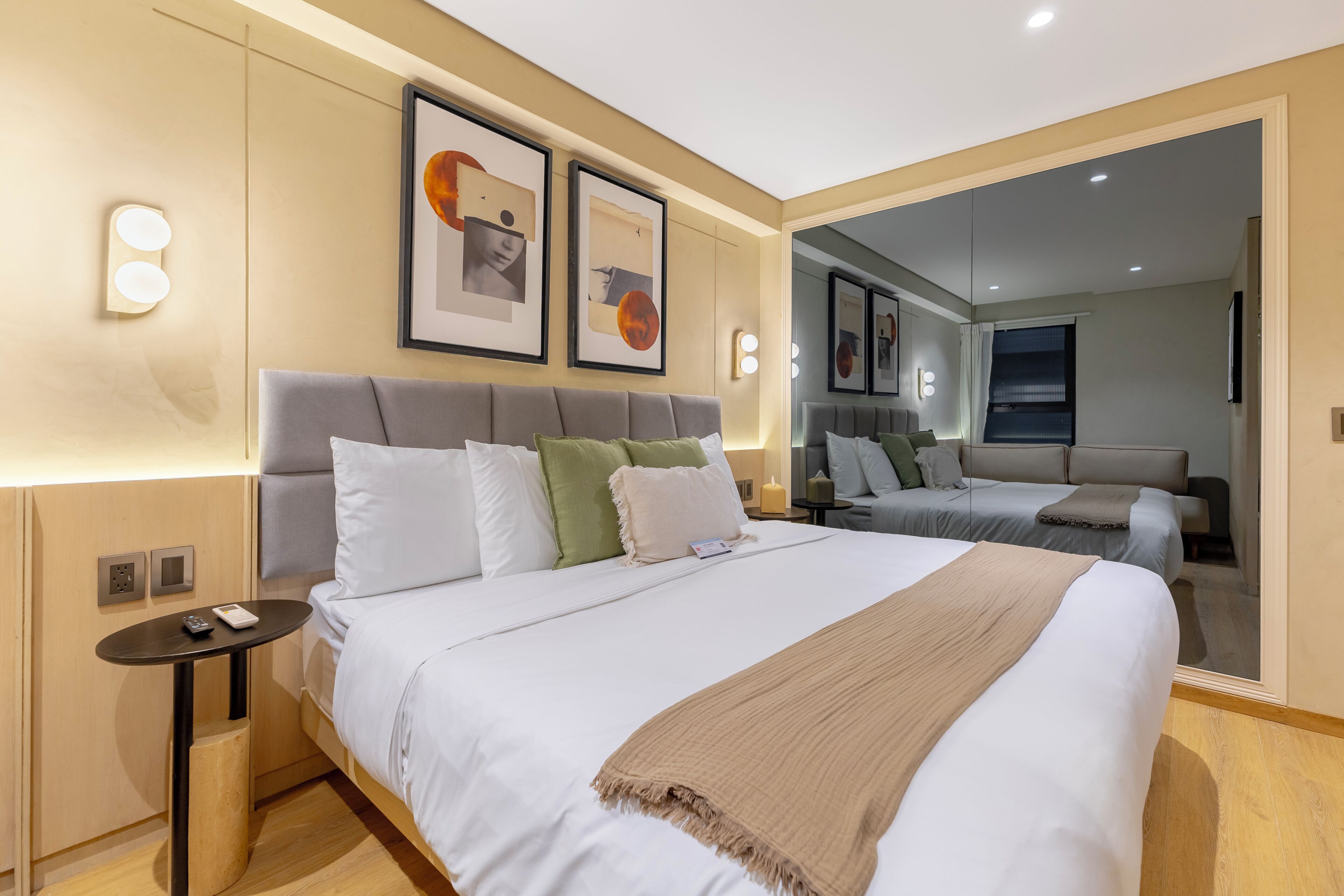 A modern hotel room with a large bed, a bedside table, a mirror, and artwork on the walls.