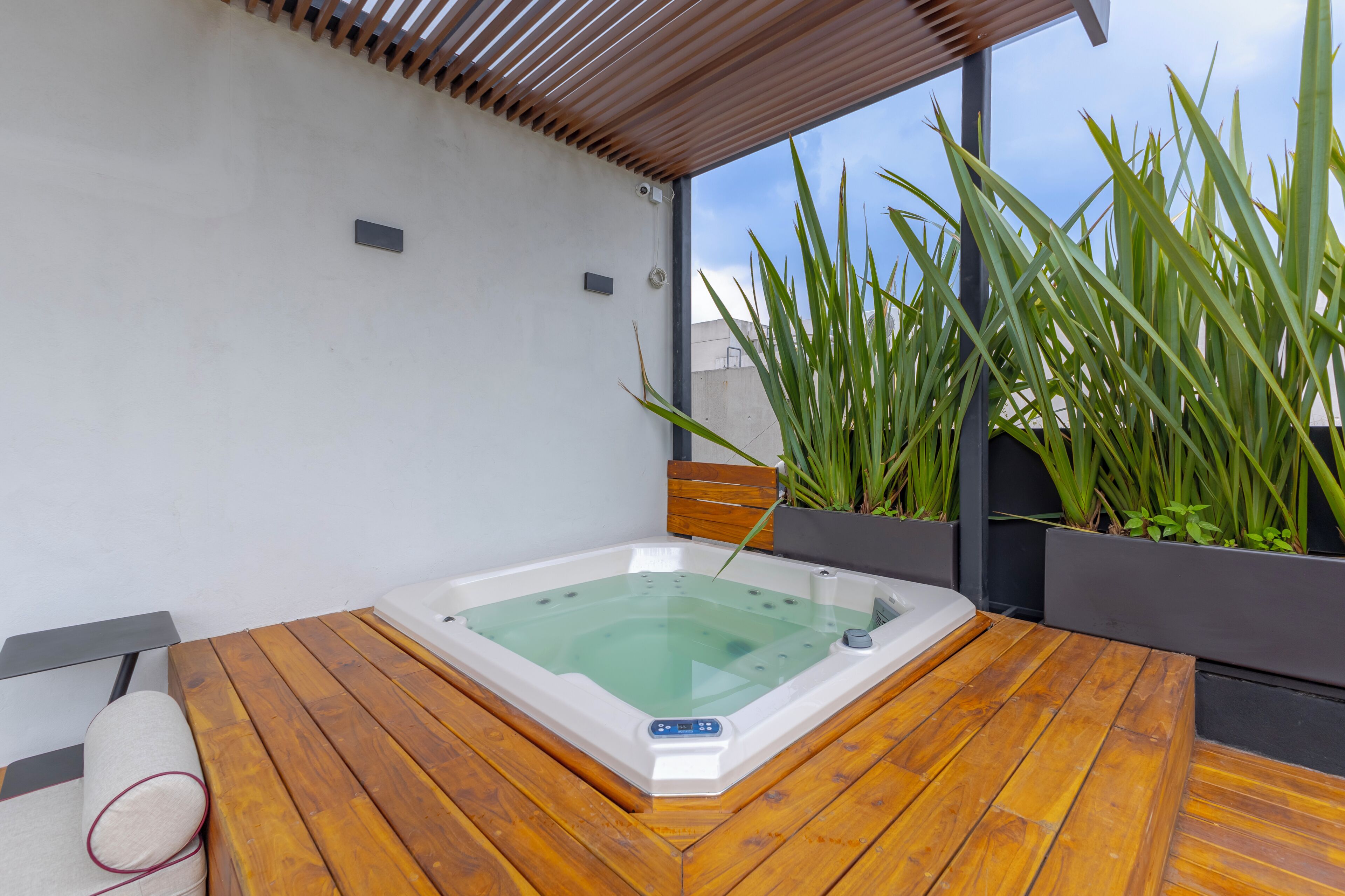 A hot tub with a wooden deck, surrounded by tall green plants and a clear sky.