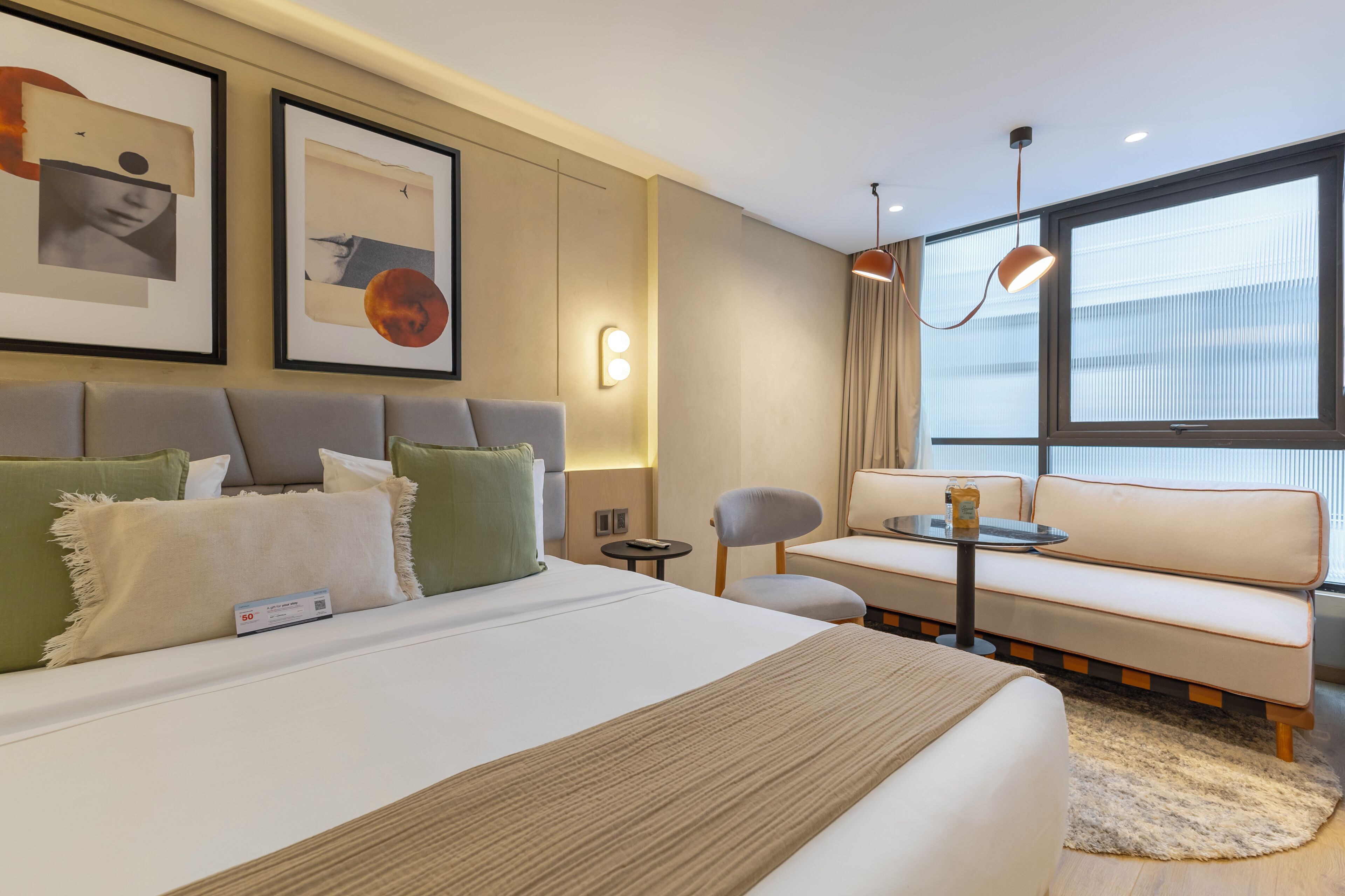 Una habitación de hotel moderna con cama, sofá, una mesita y una silla. Se observan cuadros enmarcados en la pared y una ventana con cortinas.