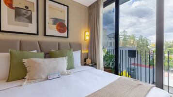 A hotel room with a large bed, a nightstand, a lamp, and a view of the city through a balcony.
