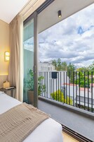 Habitación de hotel con una cama grande, una planta en maceta, una mesita de noche y vistas de la ciudad a través de un balcón con barandilla.