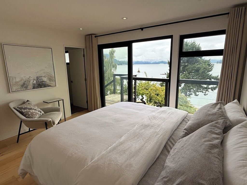 A bedroom with a large bed, a chair, and a view of a lake through large windows.