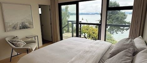 A bedroom with a large bed, a chair, and a view of a lake through large windows.