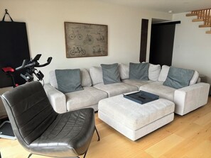 Ein modernes Wohnzimmer mit einem grauen Ecksofa, einem schwarzen Ledersessel und einem kleinen Hocker. An der Wand hängt ein fahrradbezogenes Kunstwerk, und in der Ecke steht ein schwarzes Heimtrainer.