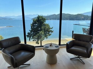 Ein Zimmer mit zwei Ledersesseln, einem kleinen runden Tisch und einem großen Fenster mit Blick auf einen See und ferne Berge.