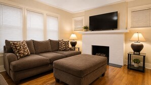 A living room with a sectional sofa, ottoman, fireplace, TV, and two lamps. - Spacious 4-bedroom apartment in fabulous Dearborn with AC (Dearborn)