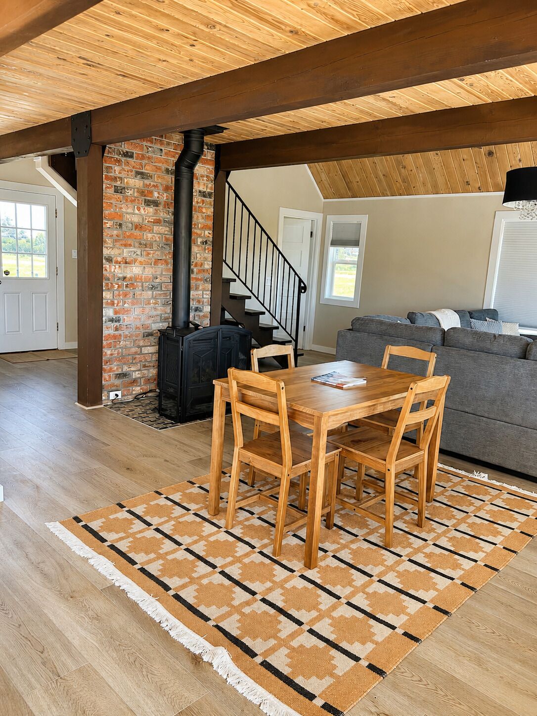 A loft-style living space with exposed wooden beams, a brick wall, a fireplace, a dining area with a table and chairs, and a gray sofa.