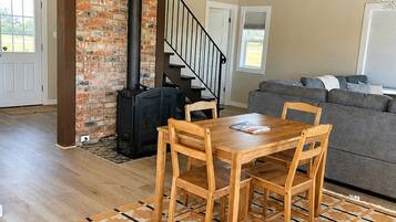 A loft-style living space with exposed wooden beams, a brick wall, a fireplace, a dining area with a table and chairs, and a gray sofa.