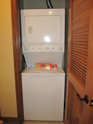 A compact, white front-loading washer and dryer unit with control knobs and a control panel, placed in a corner with a wooden door and a vent above. - Fab 2 Bedroom+ Private Hot Tub Sept 16-24 Valdoro Mountain Lodge Breckenridge (Breckenridge)