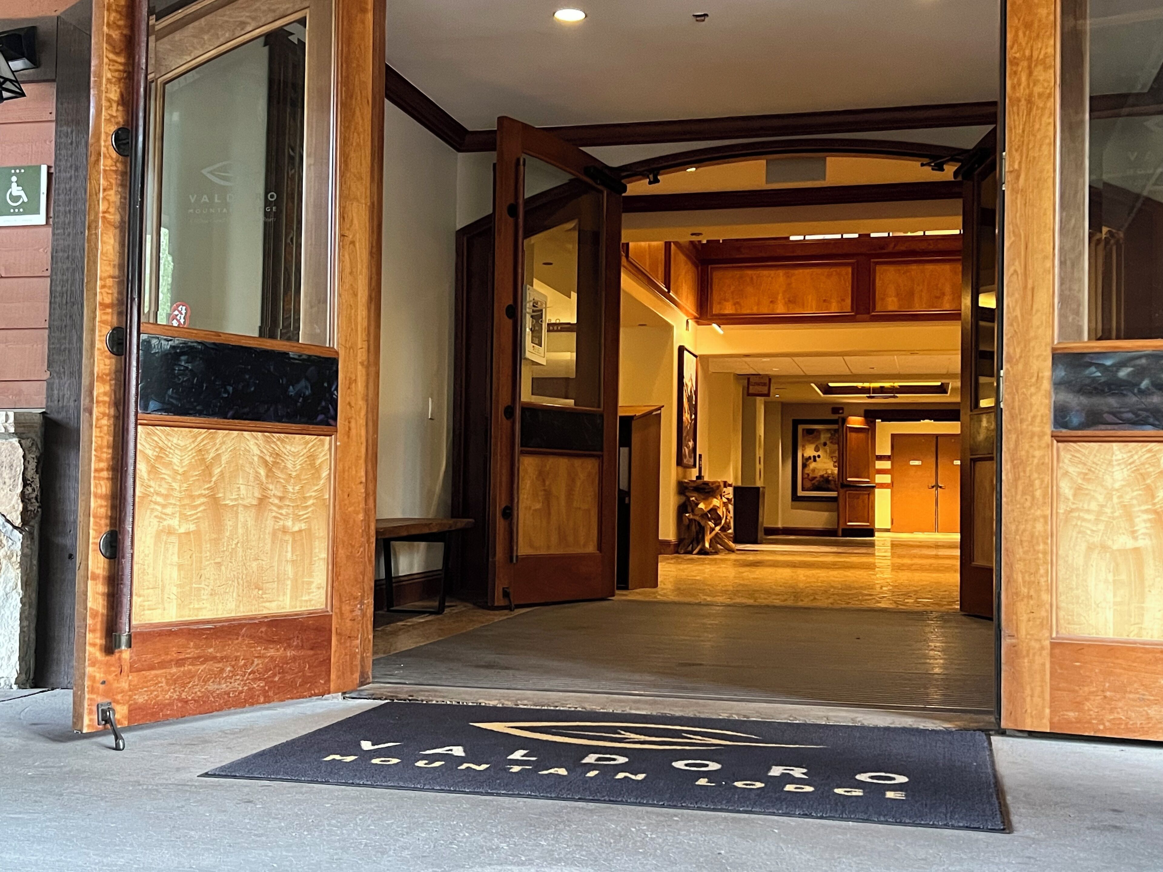 Ingresso di un hotel con arredamento in legno, un tappetino con il nome dell'hotel e un'insegna che indica l'accessibilità per disabili.
