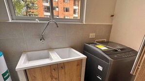 A compact kitchen with a sink, a compact washer, and a window with a view of buildings. - Premium Village 24 7 Security Balcony Fast Wifi (Medellín)