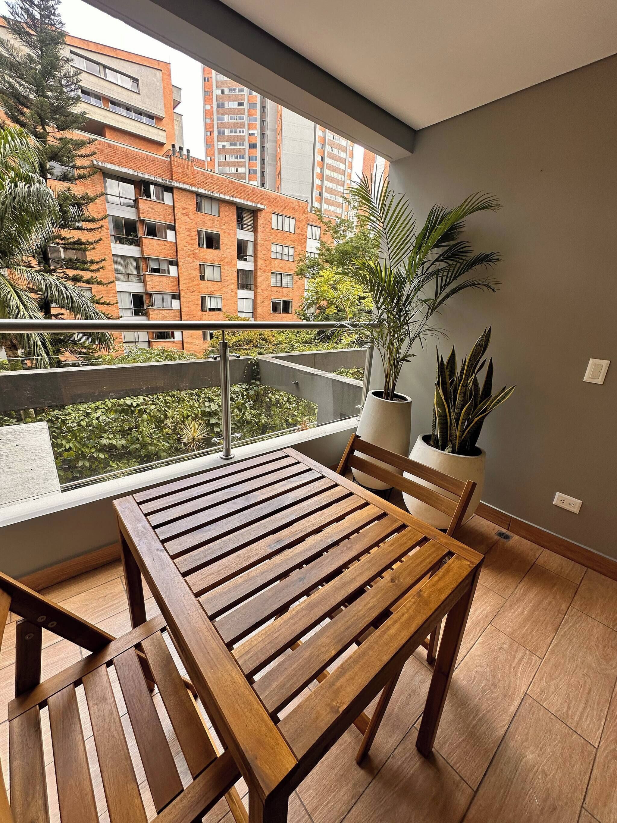 A balcony with a wooden table and chairs, potted plants, and a view of a brick building.
