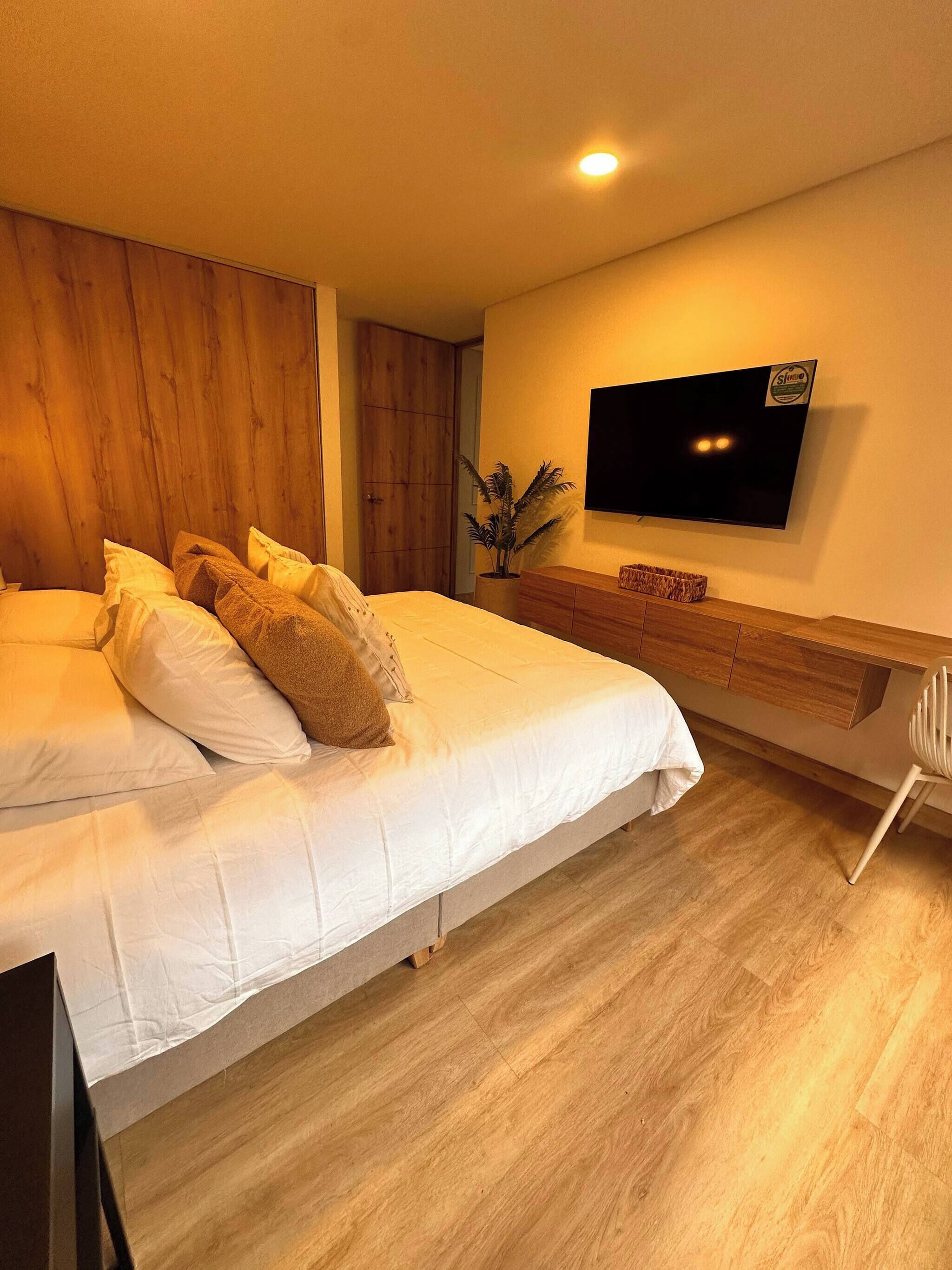 Quarto moderno com cama grande, cabeceira de madeira, televisão de tela plana na parede e cômoda de madeira.