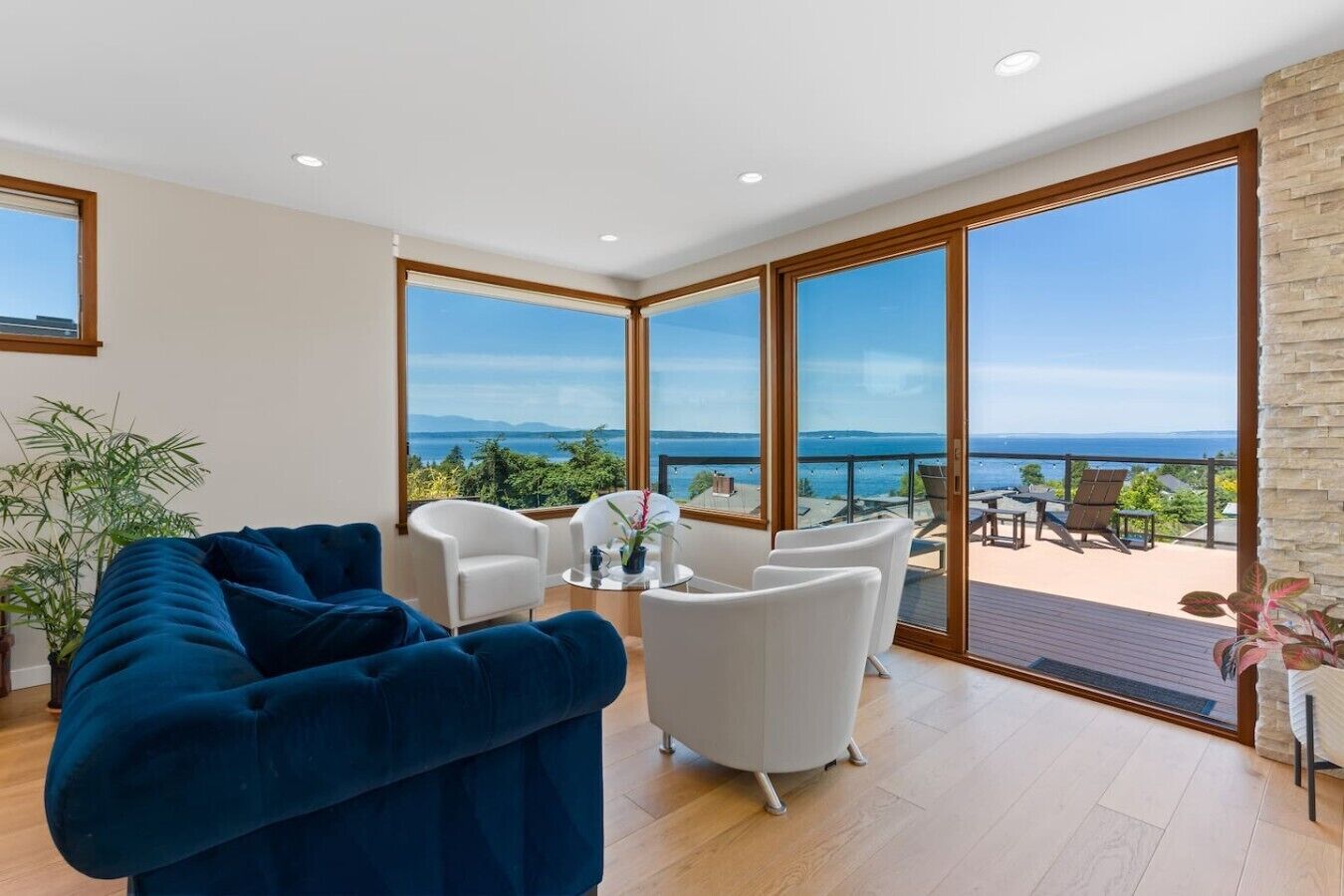 A modern living room with a sofa, chairs, a small table, and a balcony with a view of the sea.