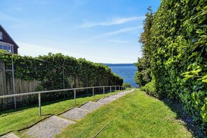 A pathway leading to a body of water, flanked by a hedge and a building.