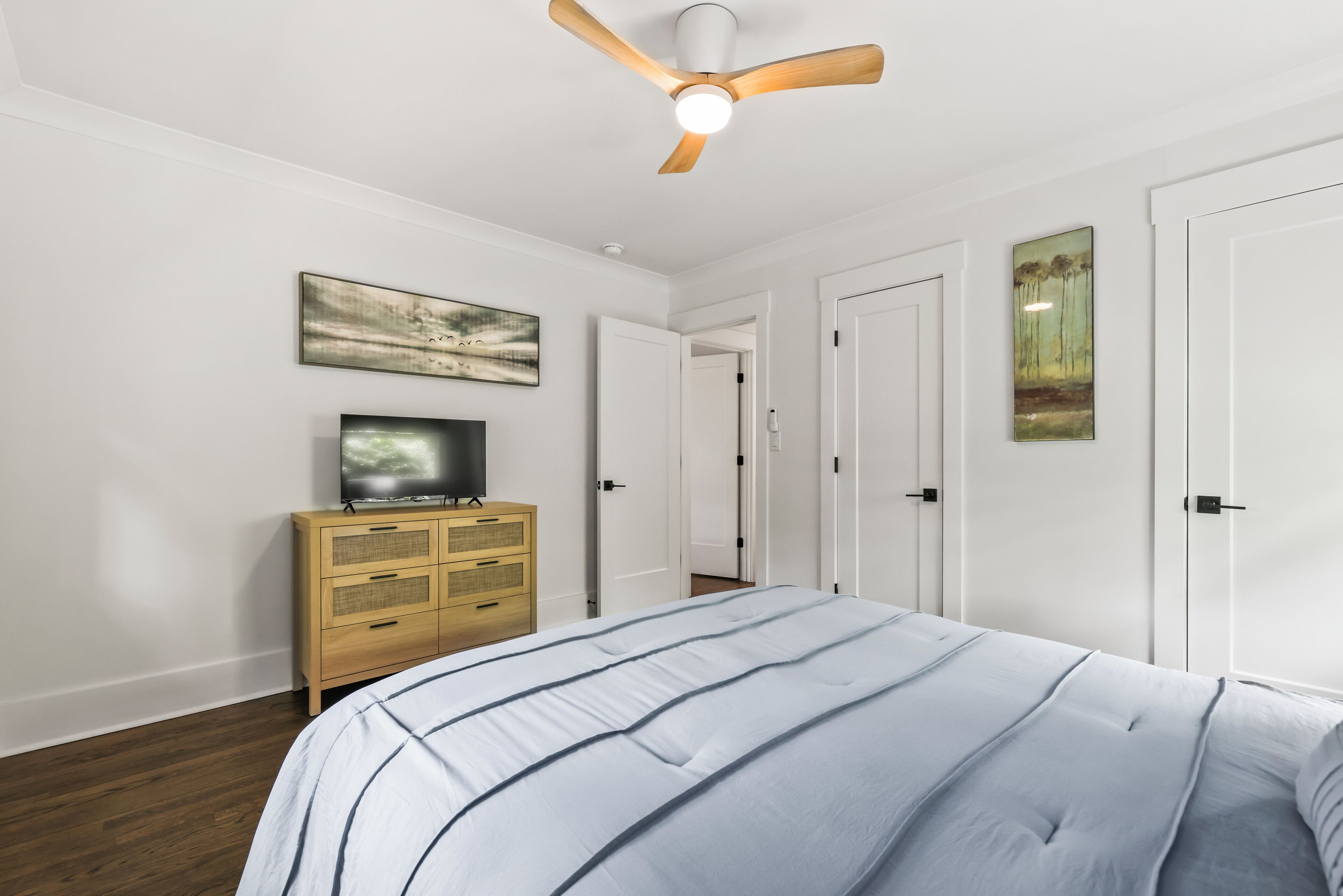A bedroom with a bed, a wooden dresser, a ceiling fan, and two doors.