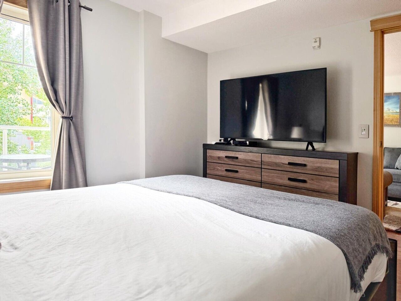 Une chambre à coucher avec un lit, un téléviseur posé sur un meuble en bois et une fenêtre avec des rideaux.