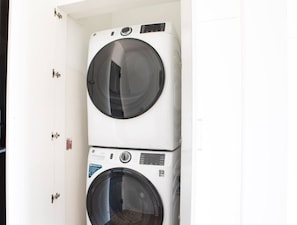 A stacked washer and dryer unit in a room with white walls and a door with a handle. - Luxury Penthouse Apt Near Brady St with Balcony and Lake View (Milwaukee)