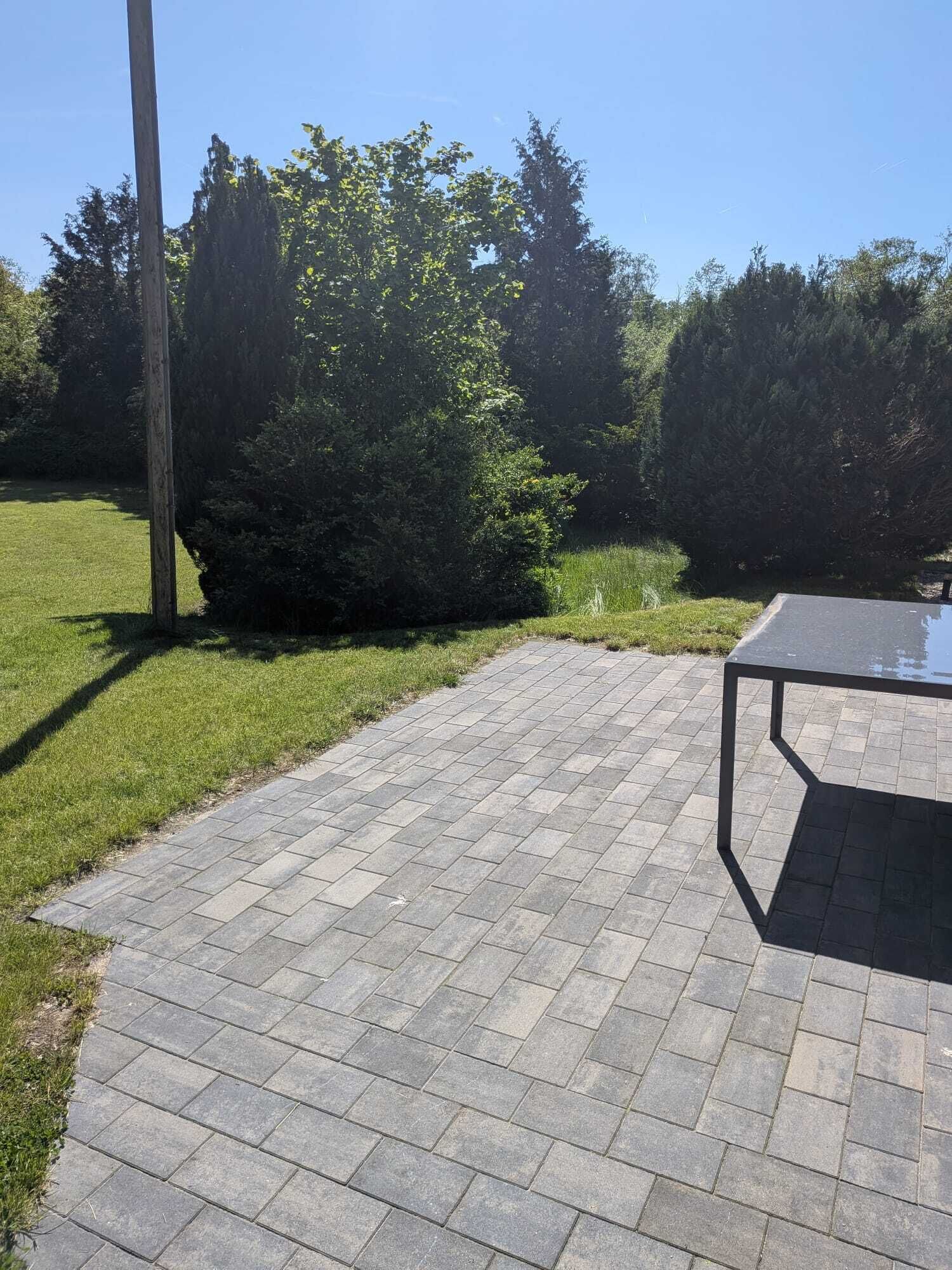 Ein gepflasterter Terrassenbereich mit Tisch und Stühlen, umgeben von Gras und Bäumen.