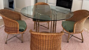 Un comedor con una mesa de vidrio rodeada de sillas de mimbre con cojines verdes, una cocina con electrodomésticos blancos y un cuadro en la pared.