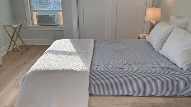 Un dormitorio con cama, ventana con persianas, mesita de noche con lámpara y una pequeña mesa con lámpara.