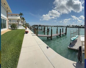 A waterfront property with a concrete walkway leading to a dock, a pool area, and a building with a white facade. - Beautiful waterfront condo with stunning views of Mooring Bay (Naples)