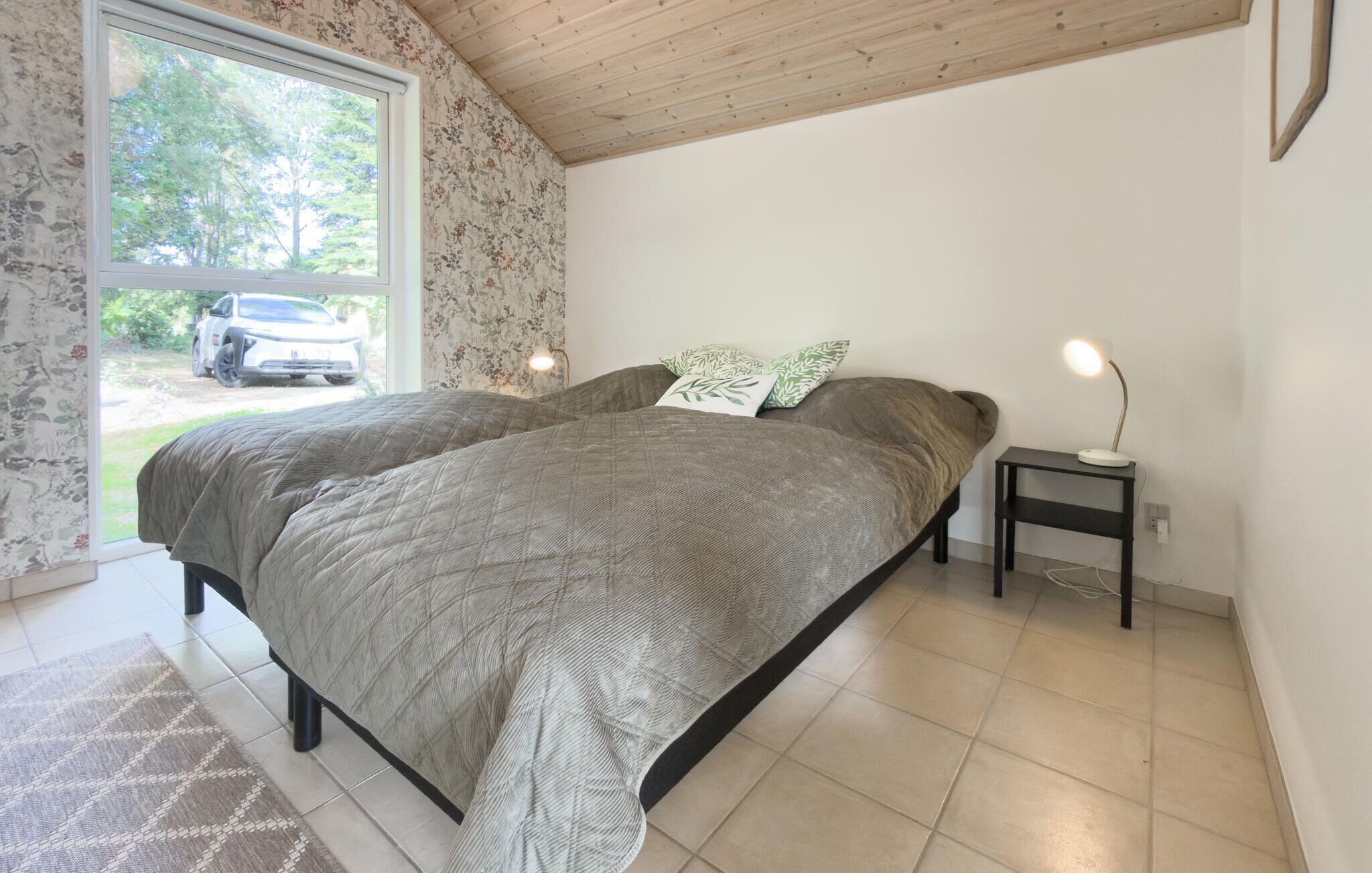 A bedroom with a bed, a bedside table with a lamp, and a window with a view of a car and greenery.