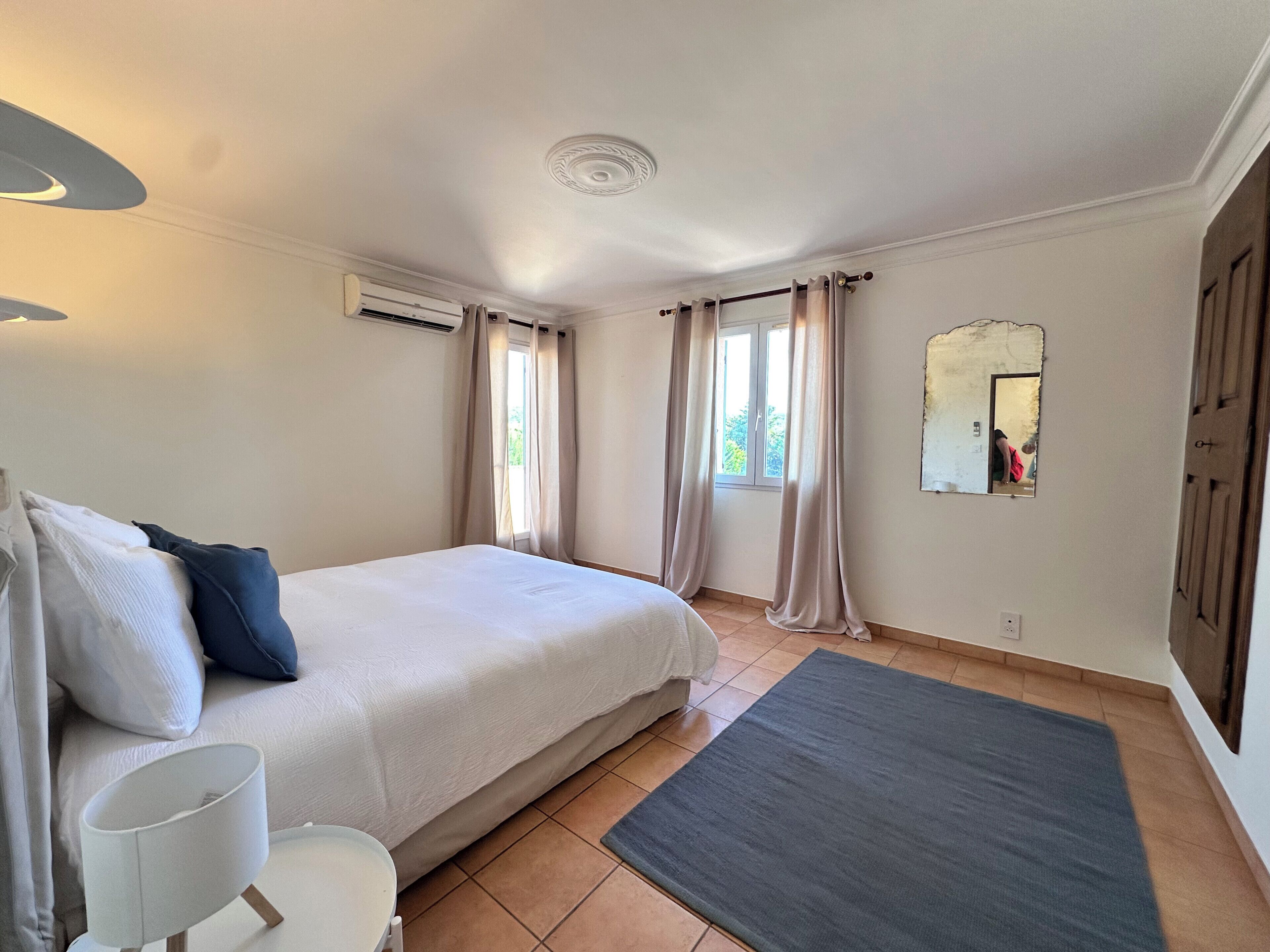 Un dormitorio con cama, mesita de noche, espejo y ventana con cortinas.