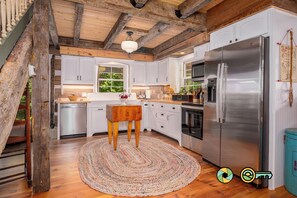 A kitchen with white cabinets, a stainless steel refrigerator, wooden beams, and a round rug. - The Sawmill Inn - pre-Civil War luxury log cabin on 70+ private acres! (Mount Jackson)
