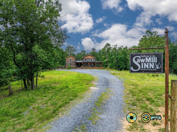 A gravel driveway leading to a cabin-style building with a porch, surrounded by trees and a clear sky.