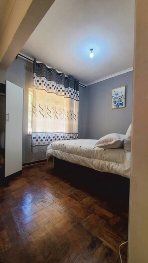 A bedroom with a wooden floor, a bed with a white and black polka dot bedspread, a window with patterned curtains, and a painting on the wall. - Guest Harven - B&B (Harare)