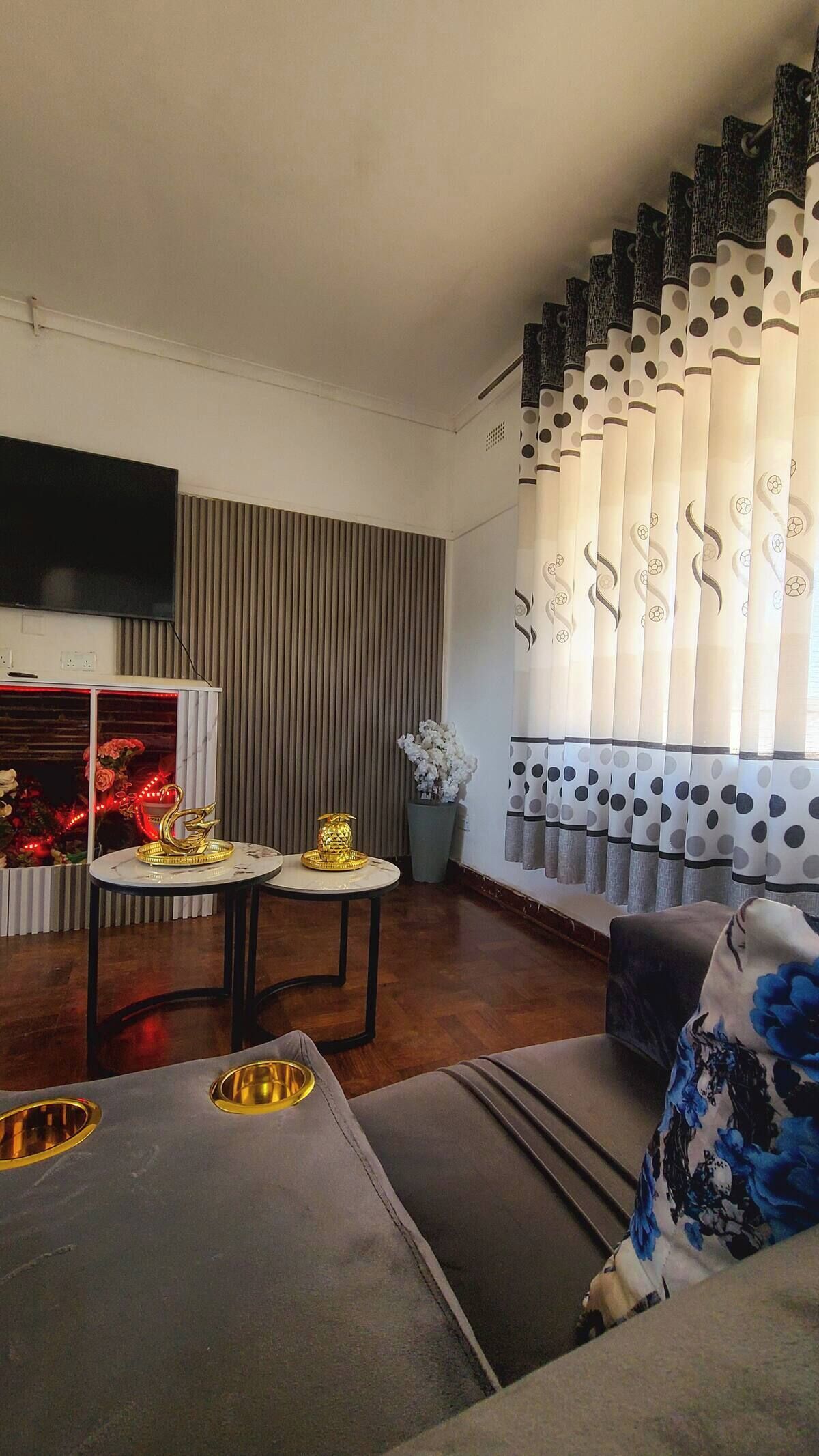 A modern living room with a flat-screen TV, a dining table with golden decorative items, a sofa with floral upholstery, and a window with patterned curtains.