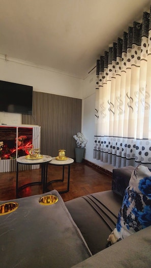 Ein modernes Wohnzimmer mit einem Flachbildfernseher, einem Esstisch mit goldenen Dekorationselementen, einer floral gepolsterten Couch und einem Fenster mit gemusterten Vorhängen.