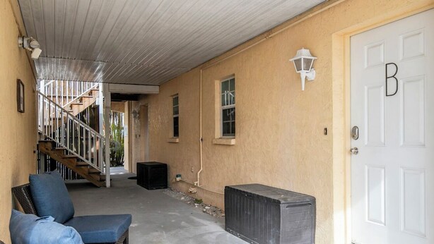 An outdoor covered patio with a staircase leading to an upper level, a white door with a letter 'B', and outdoor seating. - Snyder's Rest - A managed by Beach Retreats FL (Bradenton Beach)