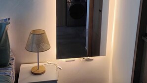 Una habitación con una pequeña mesa, una lámpara y una lavadora visible a través de un arco.
